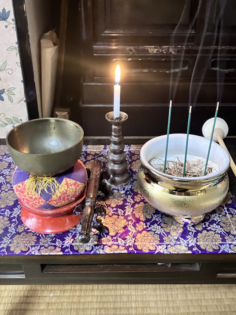 「紫色の打敷が敷かれた仏壇の上で、金色の香炉から3本の線香の煙が立ち上り、隣でロウソクの火が優しく灯っている様子。香炉の中には、日々供養されたお線香の灰が大切に積もっている。」
